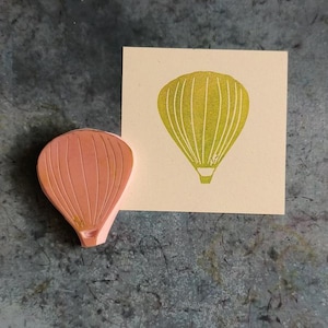 Pode incluir: Um carimbo de borracha rosa de um bal&atilde;o de ar quente com uma impress&atilde;o amarela do bal&atilde;o em um cart&atilde;o bege.