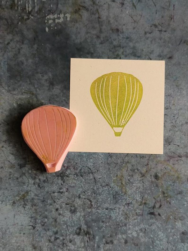 Pode incluir: Um carimbo de borracha rosa de um bal&atilde;o de ar quente com uma impress&atilde;o amarela do bal&atilde;o em um cart&atilde;o bege.