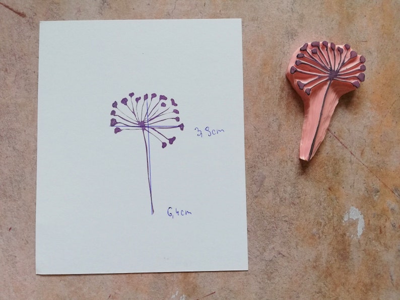 May include: A purple line drawing of a dandelion on a white sheet of paper with measurements in inches. A pink rubber stamp of a dandelion is shown to the right of the paper.