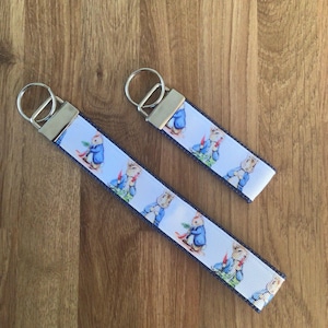 May include: Two keyrings with a white ribbon featuring a repeating pattern of a cartoon rabbit in a blue jacket. The ribbon is edged with a blue denim-like trim. Each keyring has a silver-coloured metal ring and a silver-coloured metal clasp.