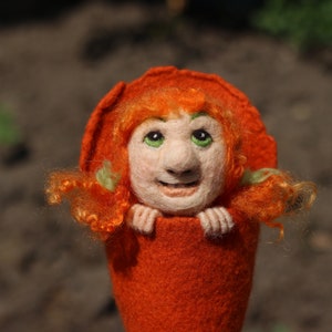 Könnte beinhalten: Handgemachte Filzpuppe mit lächelndem Gesicht, grünen Augen und orangefarbenem Haar, das aus einem orangefarbenen, kegelförmigen Körper herausragt. Die Hände der Puppe sind sichtbar, und der Hintergrund ist unscharf.