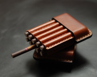May include: A brown leather cigarette case, open to reveal several cigarettes. The case is rectangular with rounded edges and a stitched design. One cigarette is partially visible, extending from the case.
