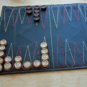 May include: A black leather backgammon board with red and yellow stitching. The board is rolled up on the right side. Wooden game pieces are arranged on the board. The game pieces are brown and beige.