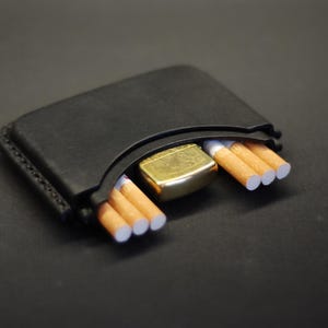 May include: A black leather case with visible stitching along the edges, designed to hold small items. The case contains a gold-toned metal lighter positioned in the center, flanked by three cigarettes on the left side and three cigarettes on the right.