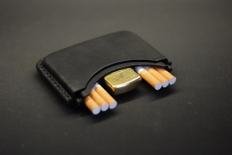May include: A black leather case with visible stitching along the edges, designed to hold small items. The case contains a gold-toned metal lighter positioned in the center, flanked by three cigarettes on the left side and three cigarettes on the right.