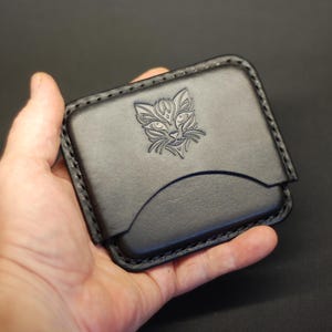 May include: A black leather card holder featuring a debossed cat face design on the front panel. The rectangular accessory has rounded corners, visible black stitching along the edges, and a curved pocket opening for storing cards or small items.
