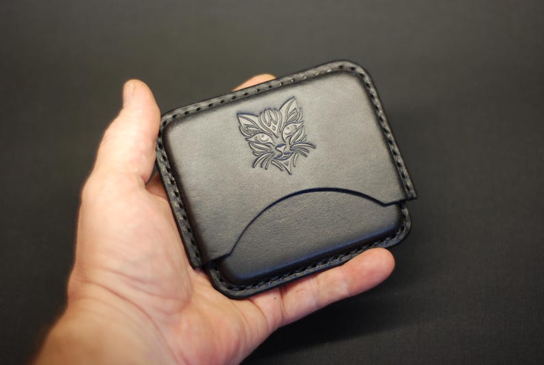 May include: A black leather card holder featuring a debossed cat face design on the front panel. The rectangular accessory has rounded corners, visible black stitching along the edges, and a curved pocket opening for storing cards or small items.