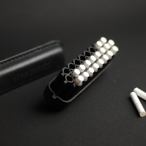 Puede incluir: Una caja negra con la palabra "STAGBILL" grabada en relieve, abierta para revelar un porta cigarrillos negro y blanco con 20 cigarrillos blancos en su interior. Dos cigarrillos blancos adicionales est&aacute;n sobre una superficie negra.