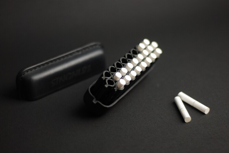 Puede incluir: Una caja negra con la palabra "STAGBILL" grabada en relieve, abierta para revelar un porta cigarrillos negro y blanco con 20 cigarrillos blancos en su interior. Dos cigarrillos blancos adicionales est&aacute;n sobre una superficie negra.