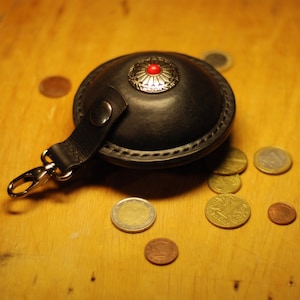 May include: A black leather coin purse with a silver and red button closure. The purse has a silver hook clasp and is surrounded by several coins.