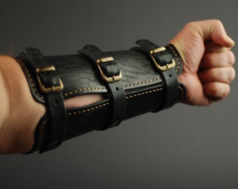 May include: Black leather arm guard with brass buckles and stitching. The guard is worn on the forearm and has a cut-out for the elbow.