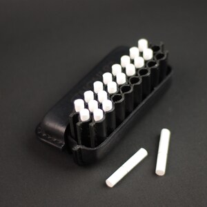 Puede incluir: Una caja negra con una fila de tizas blancas. Dos tizas adicionales est&aacute;n sobre una superficie negra.