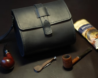 May include: A black leather tobacco pouch with a flap closure and a strap. The pouch is sitting on a dark surface next to a wooden pipe, a small pocket knife, and a pack of tobacco.