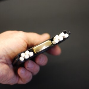 May include: A black rectangular case featuring a central gold-toned metal lighter flanked by two rows of white cylindrical sticks. The case is designed to hold the lighter securely in the middle while providing storage for the white sticks on both sides.