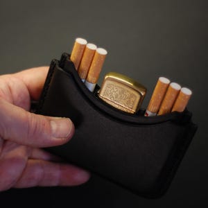 May include: A black leather case designed to hold cigarettes and a lighter. The case contains three cigarettes on the left side, three cigarettes on the right side, and a gold-toned metal lighter with an engraved floral pattern in the center.
