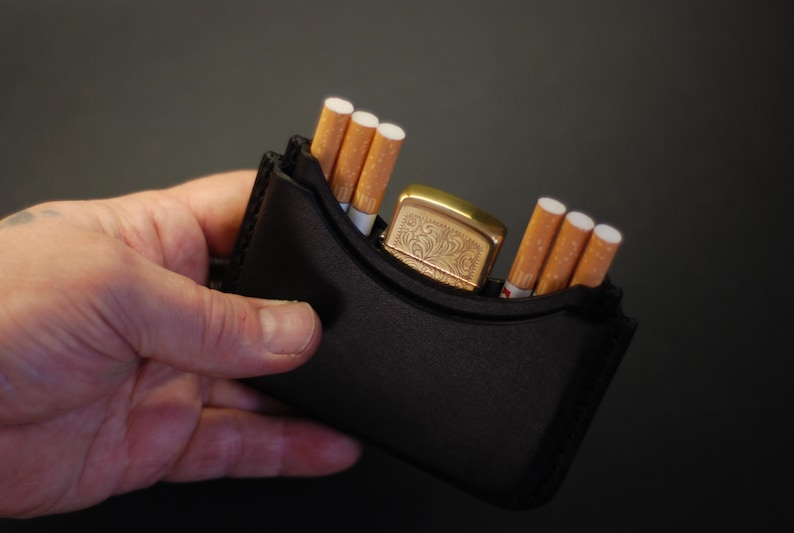 May include: A black leather case designed to hold cigarettes and a lighter. The case contains three cigarettes on the left side, three cigarettes on the right side, and a gold-toned metal lighter with an engraved floral pattern in the center.