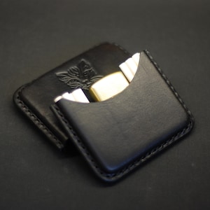 May include: A black leather card holder with visible stitching along the edges. The front features a curved cutout pocket containing a gold-toned metal lighter. A small embossed logo of a stylized animal head is visible on the back section.