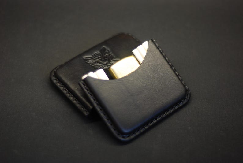 May include: A black leather card holder with visible stitching along the edges. The front features a curved cutout pocket containing a gold-toned metal lighter. A small embossed logo of a stylized animal head is visible on the back section.