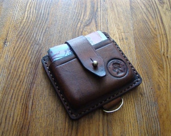 Puede incluir: Una cartera de cuero marrón con cierre de solapa y un broche de presión de latón. La cartera tiene un borde cosido y un pequeño lazo de latón en la parte inferior.