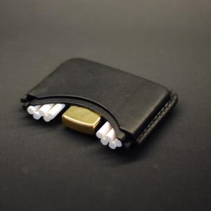 May include: A black leather case with a curved top edge, featuring a gold-toned metal lighter nestled in the center and small white cylindrical objects arranged in two groups on either side of the lighter within the leather holder.