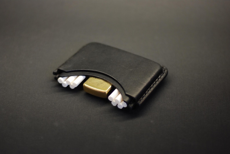 May include: A black leather case with a curved top edge, featuring a gold-toned metal lighter nestled in the center and small white cylindrical objects arranged in two groups on either side of the lighter within the leather holder.
