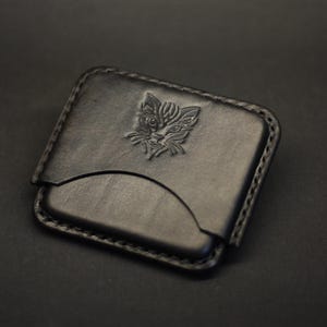 May include: A black leather card holder featuring a rectangular shape with rounded corners and visible stitching along the edges. The front surface displays a debossed graphic of a stylized cat face centered in the upper portion of the leather.