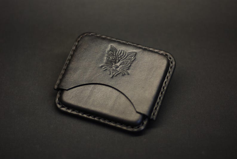 May include: A black leather card holder featuring a rectangular shape with rounded corners and visible stitching along the edges. The front surface displays a debossed graphic of a stylized cat face centered in the upper portion of the leather.