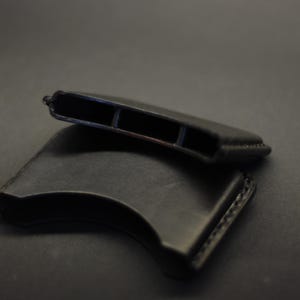 May include: Two black leather sleeves with a curved, crescent-like shape. Each sleeve features visible stitching along the edges and an open, hollow interior compartment designed to hold items securely within its structured, smooth leather frame.