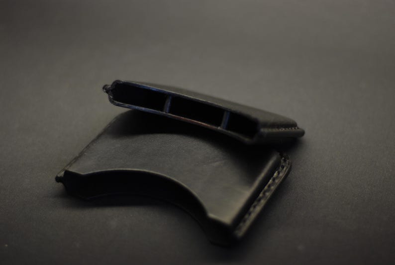 May include: Two black leather sleeves with a curved, crescent-like shape. Each sleeve features visible stitching along the edges and an open, hollow interior compartment designed to hold items securely within its structured, smooth leather frame.