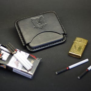 May include: A black leather cigarette case featuring an embossed cat head design, a pack of cigarettes with black filters and white bodies, a gold-toned rectangular metal lighter with an engraved pattern, and two loose cigarettes with black filters.