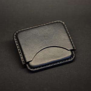 May include: A black leather card holder with rounded corners and visible stitching along the edges. The front features a curved cutout design for easy access to cards, showcasing a smooth, matte finish on the surface of the leather material.