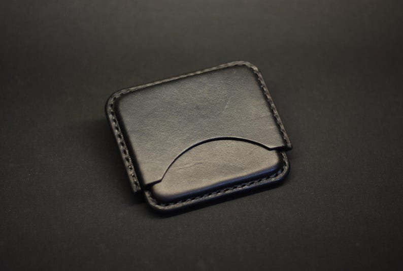 May include: A black leather card holder with rounded corners and visible stitching along the edges. The front features a curved cutout design for easy access to cards, showcasing a smooth, matte finish on the surface of the leather material.