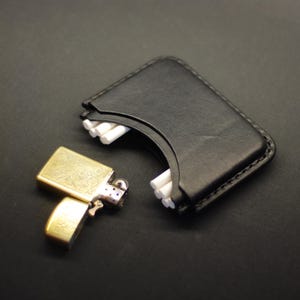 May include: A black leather cigarette case with a curved opening revealing white cigarettes, paired with a gold-toned metal flip-top lighter featuring an intricate engraved floral pattern on its rectangular surface and a hinged lid mechanism.