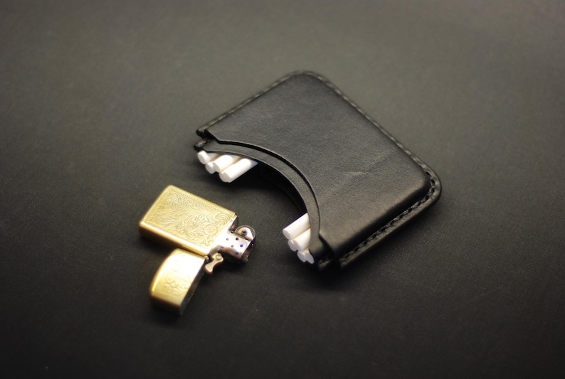 May include: A black leather cigarette case with a curved opening revealing white cigarettes, paired with a gold-toned metal flip-top lighter featuring an intricate engraved floral pattern on its rectangular surface and a hinged lid mechanism.