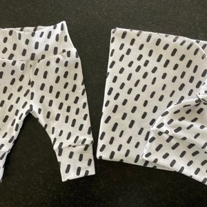 Könnte beinhalten: Ein Set Babykleidung in einem weißen und schwarzen Muster. Das Set beinhaltet eine Leggings und eine Mütze. Die Leggings hat einen schwarzen elastischen Bund und die Mütze hat einen Knoten oben.