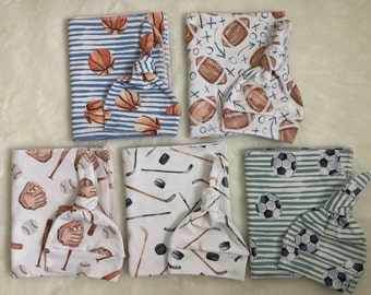 May include: Set of five baby swaddles and matching hats featuring different sports themes. The swaddles are white with various sports-themed prints, including baseball, hockey, football, soccer, and basketball. The hats are made of a soft, stretchy fabric and are tied with a knot at the top.