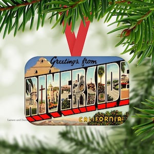 May include: Rectangular Christmas ornament featuring a vintage postcard design with the words "Greetings from Riverside" and "California." The ornament has a red ribbon for hanging and is set against a backdrop of evergreen branches.