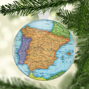 May include: A round, blue and white Christmas ornament with a map of Spain and Portugal on it. The ornament is hanging from a pine tree branch.