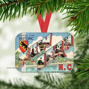 May include: Rectangular Christmas ornament featuring a vintage postcard design with the text "Greetings from Historic New Bern N.C." The ornament has a red ribbon for hanging and is surrounded by green pine needles.
