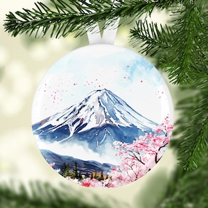 May include: A white round Christmas ornament with a watercolor painting of Mount Fuji and cherry blossoms. The mountain is blue and white with snow on the top. The cherry blossoms are pink and white.