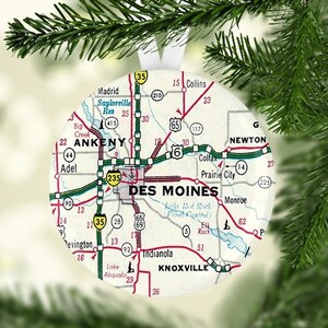 May include: A white and green Christmas ornament featuring a vintage map of Des Moines, Iowa. The map shows major roads and highways, including Interstate 35, Interstate 6, and Interstate 80. The ornament also includes the names of nearby towns, such as Ankeny, Adel, and Newton.
