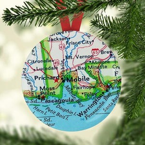 May include: A round, colorful map ornament featuring the city of Mobile, Alabama. The map shows surrounding towns and cities, including Pascagoula, Fairhope, and Pensacola. The ornament is a great way to show your pride in your hometown or to remember a special trip.