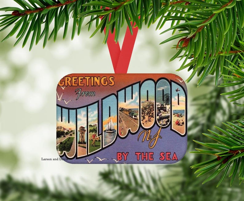 May include: Rectangular Christmas ornament with a vintage postcard design. The ornament reads "GREETINGS from WILDWOOD NJ BY THE SEA" in a stylized font, with images of a beach and boardwalk. It has a red ribbon for hanging.