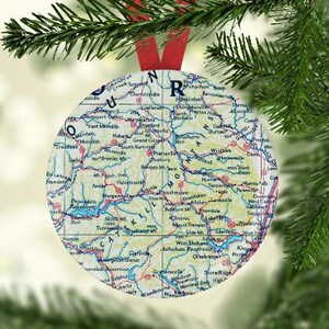 May include: A round Christmas ornament featuring a map design with red, blue, and green details. The ornament is hanging from a red ribbon and is surrounded by green pine branches.