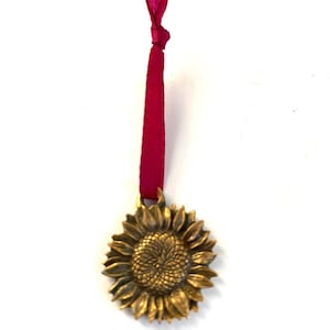 May include: A gold-colored sunflower pendant with detailed petals and a textured center. It hangs from a knotted, deep red ribbon. The pendant is likely made of metal and is designed to be hung as an ornament.