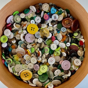 May include: A terracotta bowl overflowing with a diverse collection of buttons. The buttons vary in size, shape, and color, including yellow, green, white, and brown. Some buttons have four holes, while others have two or are heart-shaped.