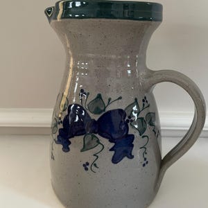 May include: A ceramic pitcher with a gray speckled body, a dark green rim, and a curved handle. The pitcher is decorated with blue bow and leaf designs. The pitcher is approximately 9 inches tall.