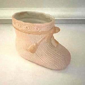 May include: A pale pink ceramic boot-shaped planter featuring a textured knit pattern on the exterior. The top rim is decorated with a raised circular motif and a sculpted rope detail that ties into a bow on the side of the boot.