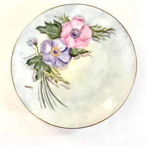 May include: A round, decorative plate with a gold rim. The plate features hand-painted flowers in shades of pink and purple, with green leaves. The background is a soft, pale green. The artist's signature, "Woolley", is visible.