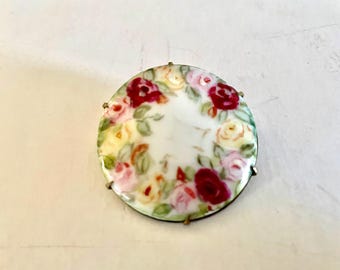 May include: A white porcelain brooch with a floral design. The brooch features a circle of red, pink, and yellow roses with green leaves on a white background. The brooch is accented with gold-colored metal.